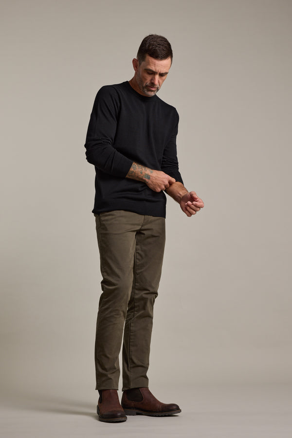 A man in a black sweater and Barkers Carnaby Chino in olive green stands against a plain background, looking down while adjusting his sleeve. He completes the look with brown boots.