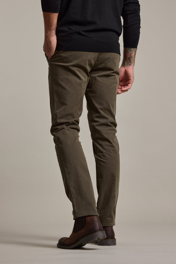 Someone in Barkers Carnaby Chino (olive green), a black sweater, and brown boots stands with their back to the camera, one hand in their pocket, against a plain light background.