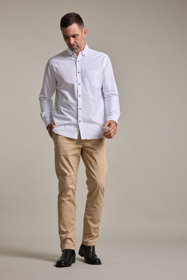 A man stands against a plain background wearing Barkers' Carnaby Chino pants in beige, paired with a white button-up shirt and black dress shoes. Made from responsibly sourced cotton, he looks down with his hands by his sides.