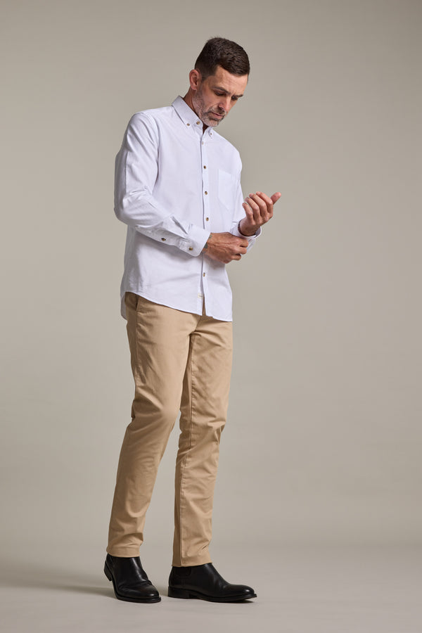 A man stands against a plain background, looking down as he buttons the cuff of his white dress shirt. He is wearing Barkers Carnaby Chino pants made from responsibly sourced cotton and black shoes.