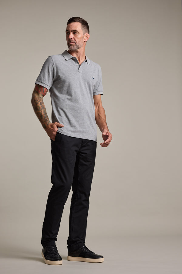 A man with short hair and tattoos on his left arm stands against a plain background, wearing Barkers Dundas Chino trousers with a gray polo shirt and black sneakers with white soles. He looks to his left, expression neutral.