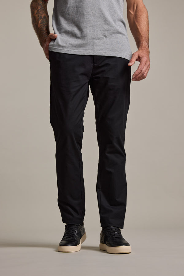 Wearing Barkers Dundas Chino trousers in black and black sneakers with white soles, a person stands hands-in-pockets against a plain background. Only the relaxed fit lower half is visible, paired with a light gray t-shirt.