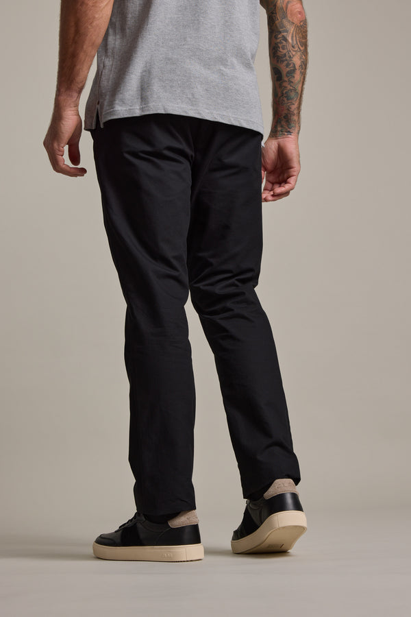 A person stands facing away, wearing a gray t-shirt, Barkers Dundas Chino trousers in black, and black sneakers with white soles; a tattoo is visible on their left arm.
