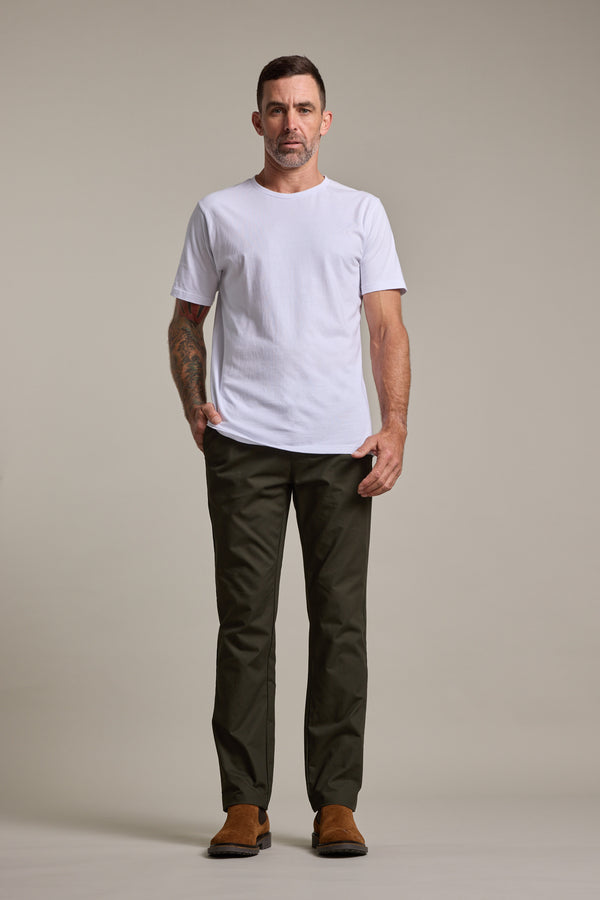 A man stands facing forward with a neutral expression, wearing a plain white t-shirt, Barkers Dundas Chino trousers in dark green, and brown shoes, one hand in his pocket against a plain light gray background.