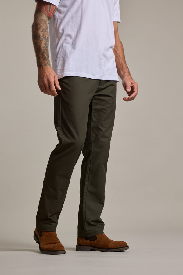 A man stands against a plain background wearing a white t-shirt, Barkers Dundas Chino in relaxed-fit olive green, and brown suede boots. His left arm is tattooed, and his head is out of frame.