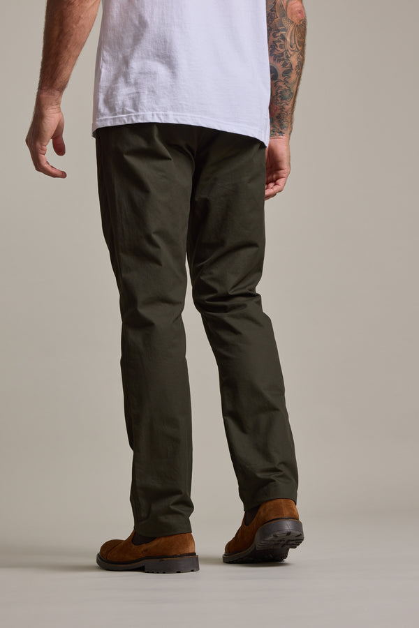 A person faces away, wearing a white T-shirt, Barkers Dundas Chino cotton twill trousers in dark olive, and brown shoes. Their left arm is tattooed. The background is plain and neutral.