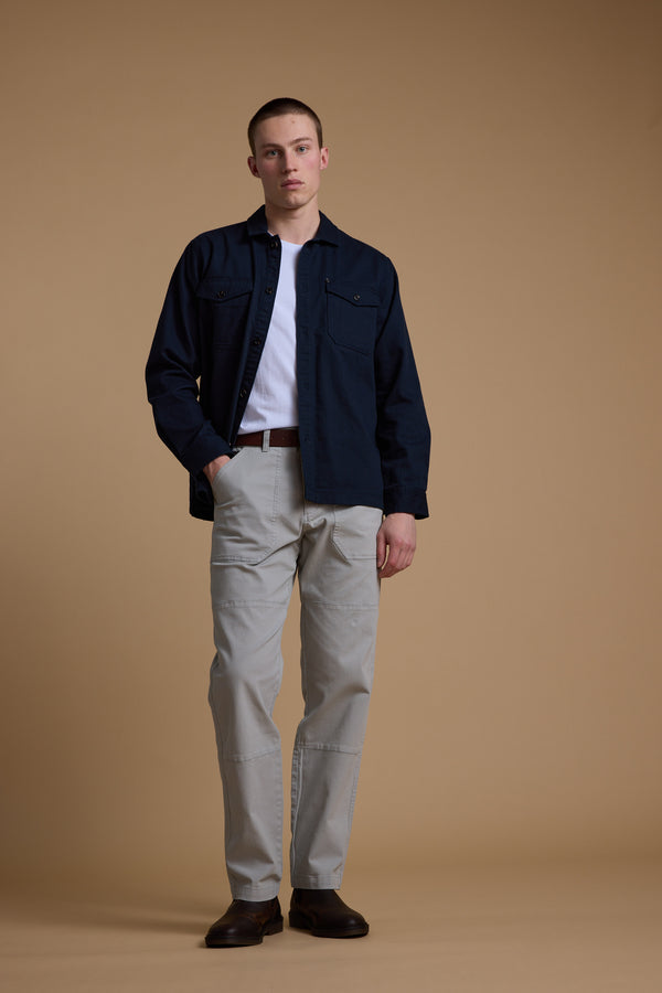 A man stands against a plain tan background wearing Barkers' Tidal GD Utility Pant in light gray with a tapered fit, paired with a navy button-up, white t-shirt, brown belt, and dark brown shoes. One hand is in his pocket and he has a neutral expression.
