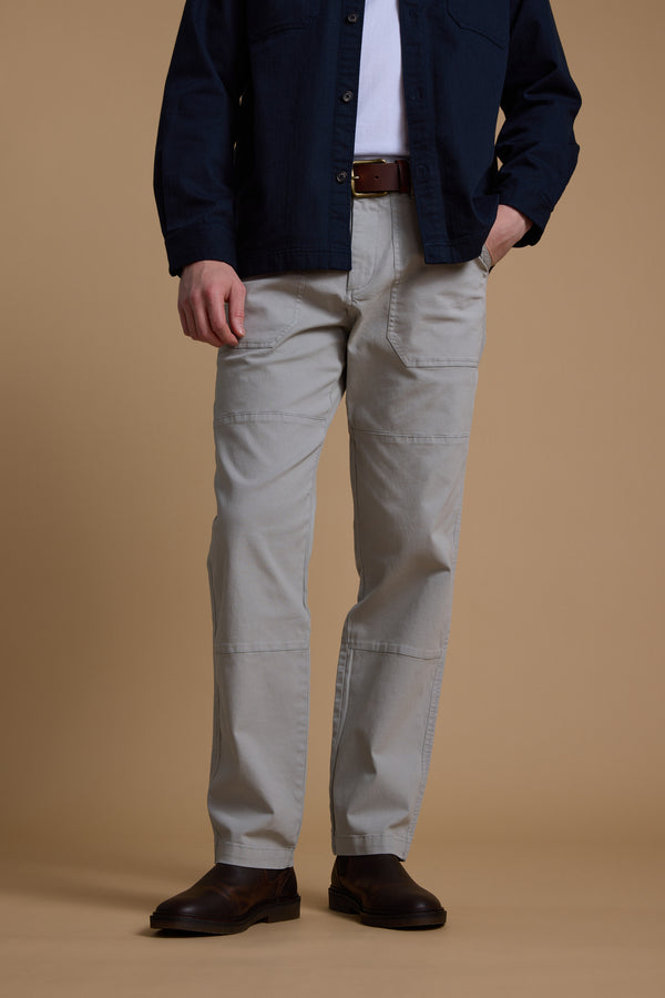 A person stands against a beige background wearing Barkers' Tidal GD Utility Pant in light gray, paired with a dark navy shirt over a white tee, a brown belt, and dark brown shoes. The person's face is not visible.
