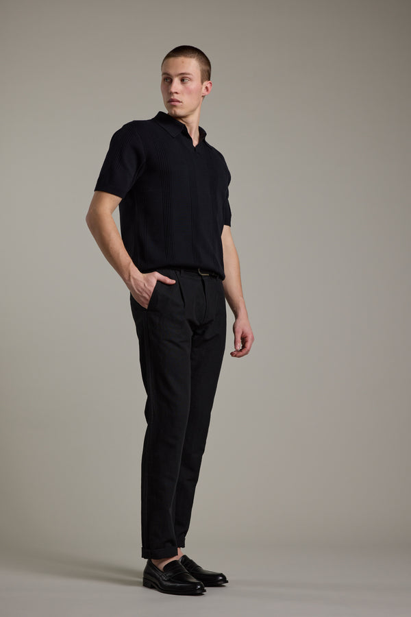 A man stands against a gray background in smart-casual style, wearing a short-sleeve black shirt, Barkers Newman Linen Pant in black, and loafers. With one hand in his pocket, he looks to the side with a neutral expression.