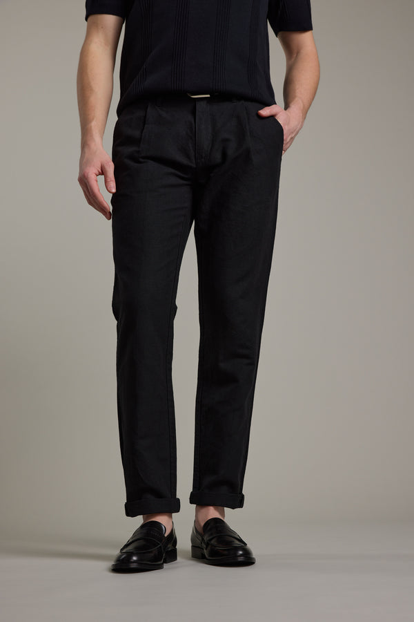 A person stands against a plain background, wearing Barkers' Newman Linen Pant in black with cuffed hems, styled smart-casually with a matching black shirt and loafers. One hand is in their pocket, the other rests relaxed by their side.