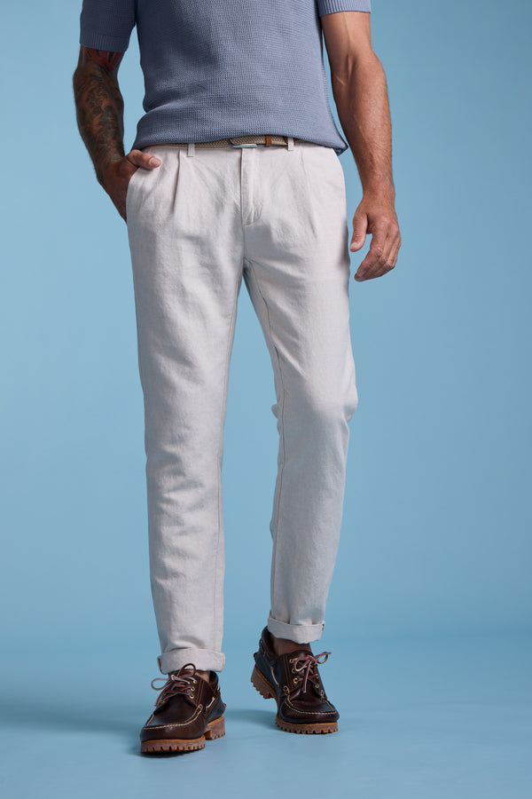 A man stands against a blue background wearing a short-sleeve blue knit shirt, Barkers' Newman Linen Pant in light beige, a brown belt, and dark brown leather shoes. A tattoo is visible on his left arm.