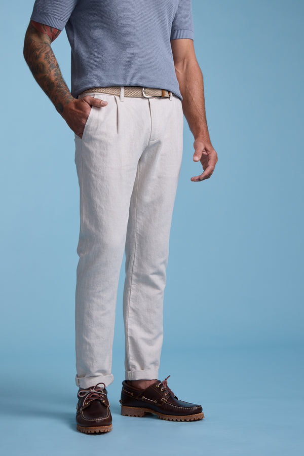 A man stands against a blue background wearing Barkers' Newman Linen Pant in beige with a belt, a short-sleeve light blue shirt, and brown leather boat shoes. One hand is in his pocket, revealing a tattoo on his left arm.