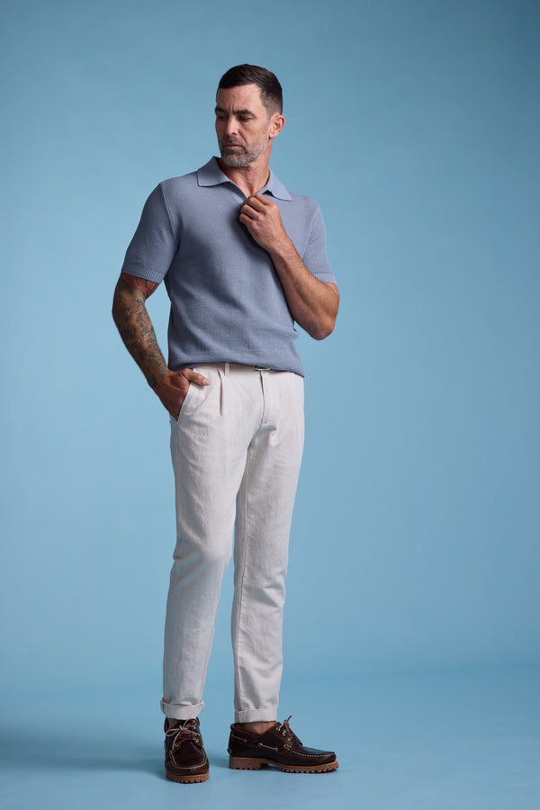 A man with short hair and a beard stands against a blue background, wearing a light blue polo, Barkers Newman Linen Pant in light beige, and brown boat shoes. One hand is in his pocket while the other adjusts his collar.