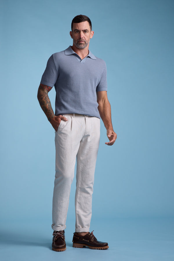 A man with short dark hair and tattoos stands against a blue background, wearing a light blue short-sleeve knit polo, Barkers' Newman Linen Pant in beige, and brown leather shoes.