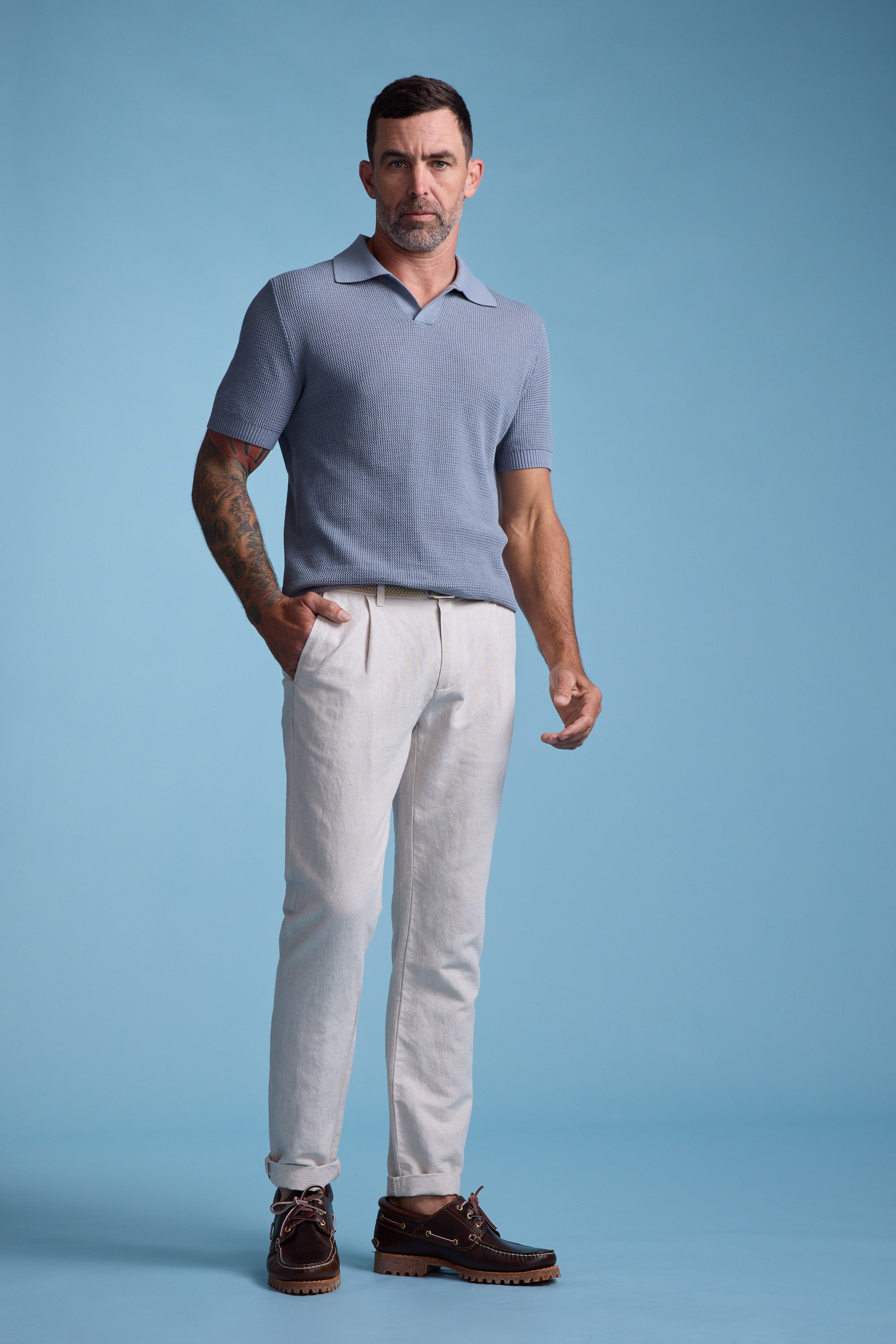 A man with short dark hair and a tattooed right arm stands against a light blue background, wearing Barkers’ Newman Linen Pant in beige, a light blue polo shirt, and brown leather shoes. He has one hand in his pocket and a neutral expression.