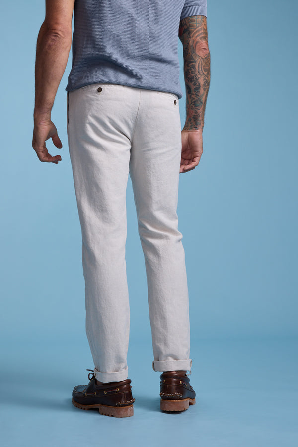 A man faces away against a blue background, wearing Barkers Newman Linen Pant—relaxed fit with buttoned back pockets—paired with a light blue short-sleeve shirt and brown leather shoes. One arm is tattooed for added style.