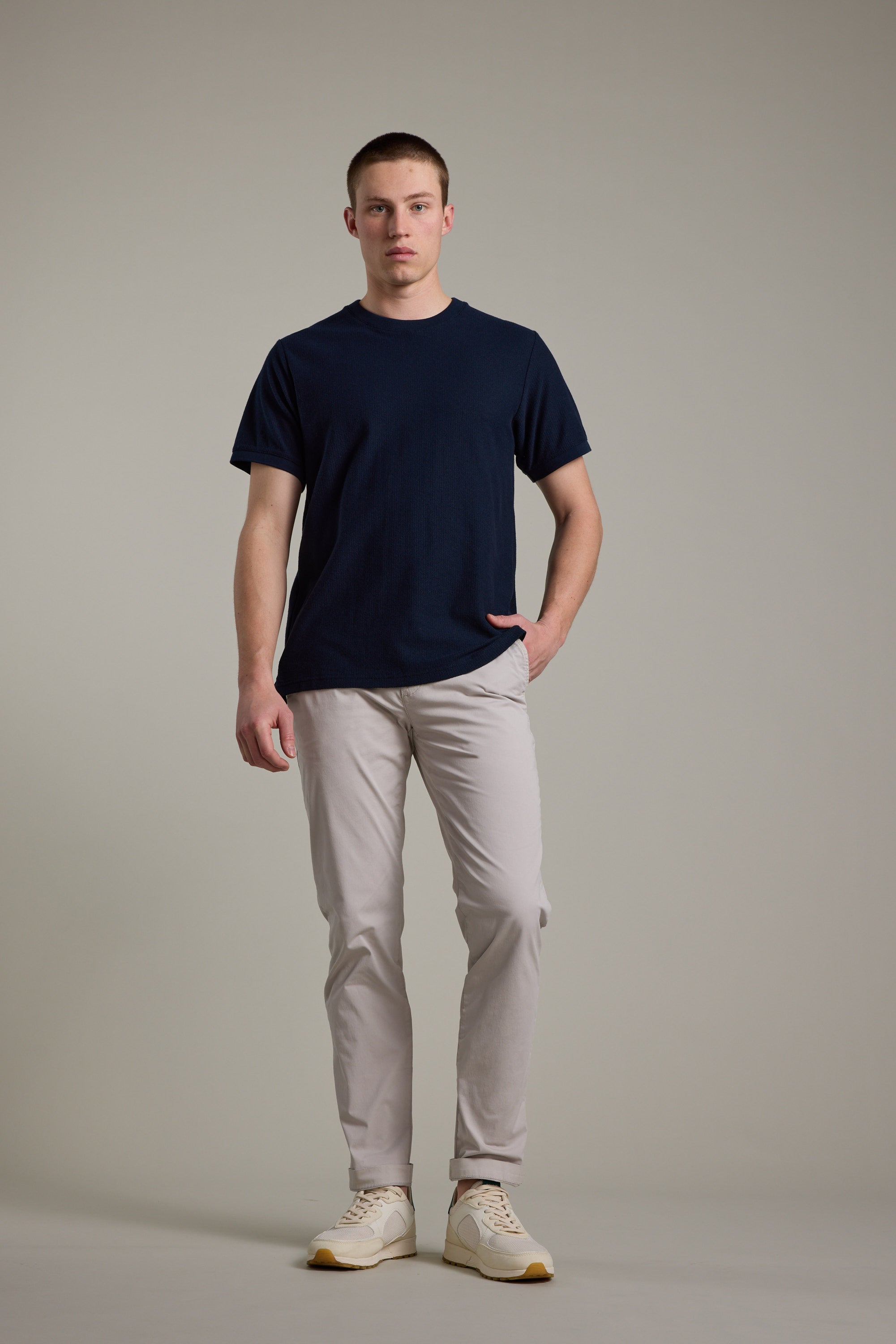 A young man stands against a neutral background, wearing Barkers Kingly Chino Pant in light beige, a navy blue t-shirt, and white sneakers. One hand is in his pocket as he looks straight ahead with a neutral expression.
