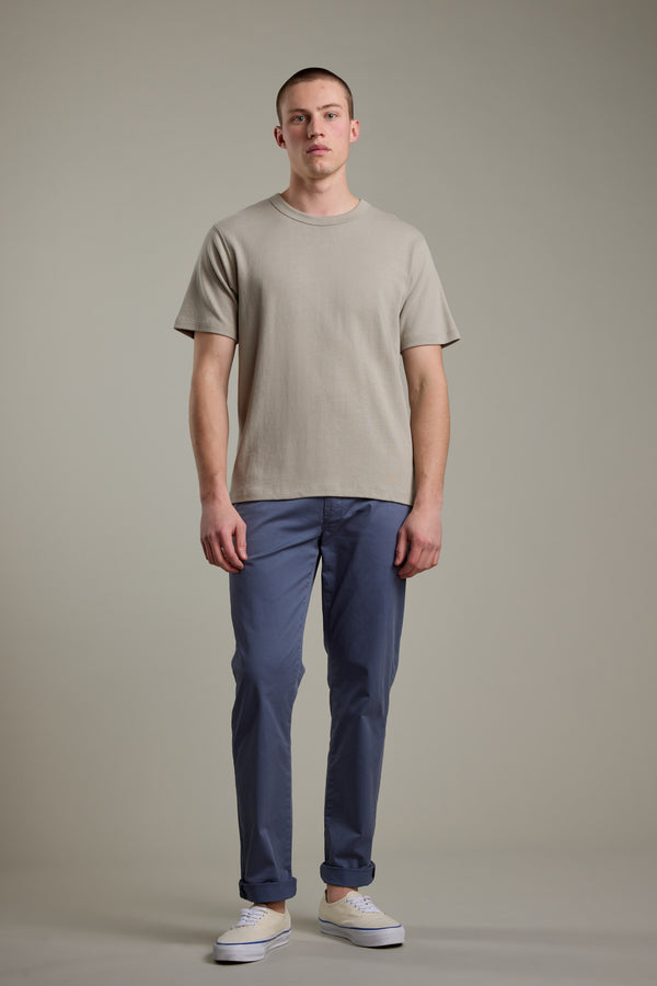 A man with short, styled hair wears a beige short-sleeve tee, Barkers Kingly Chino Pant in blue, and white sneakers against a plain background, portraying a versatile summer look with a neutral expression.