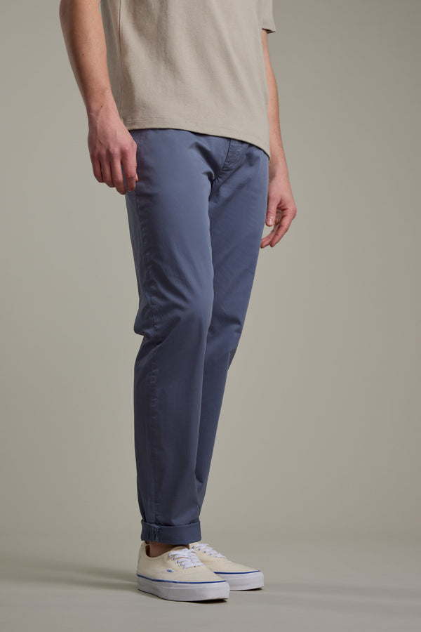 A person stands on a light gray floor wearing Barkers Kingly Chino Pant in blue, a beige t-shirt, and white sneakers. The photo is cropped at the shoulders with a plain background, highlighting these summer-ready chinos in style.
