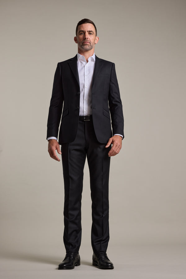 A man faces forward against a light gray background, wearing the Barkers Mayfair ZQrx Suit Jacket in sleek black, paired with a white dress shirt and black dress shoes.