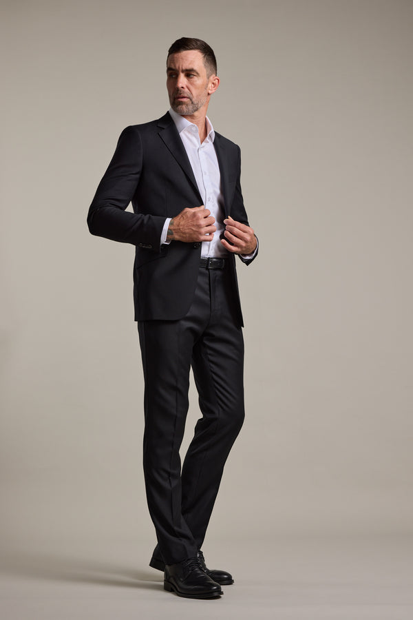 A man with short hair and a beard wears the Barkers Mayfair ZQrx Suit Jacket in classic black, made from fine merino wool. Against a plain background, he adjusts his cuff—perfect for a stylish wedding suit look.