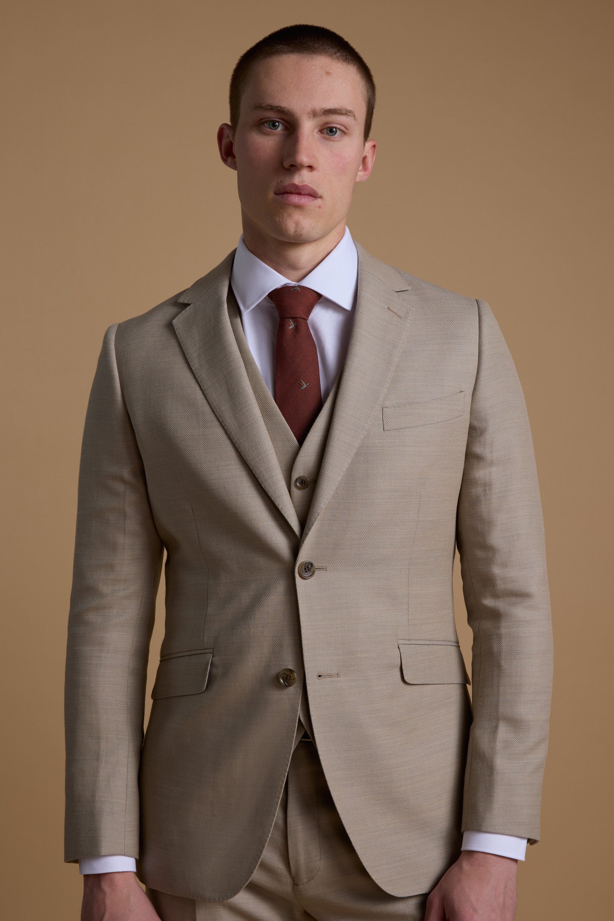 A man in the Barkers Lister Texture Suit Jacket, featuring a beige wool-linen blend, paired with a white shirt and brown tie, stands against a tan backdrop—an ideal summer wedding suit.