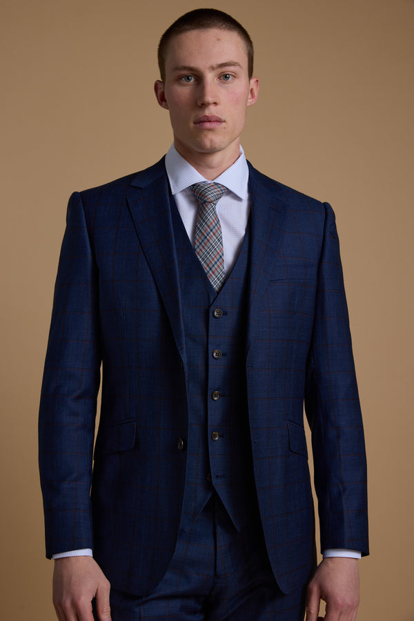 A man with short hair wears the Barkers Grafton Check Suit Jacket in dark blue, paired with a matching vest, white dress shirt, and patterned tie, standing against a plain beige background.