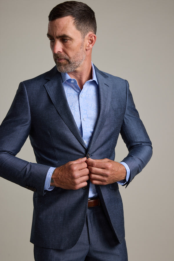 A man with short dark hair and a beard wears the Barkers Charles Texture Suit Jacket over a light blue shirt, looking left while buttoning up. A plain background highlights this modern tailoring, ideal as a summer wedding suit option.