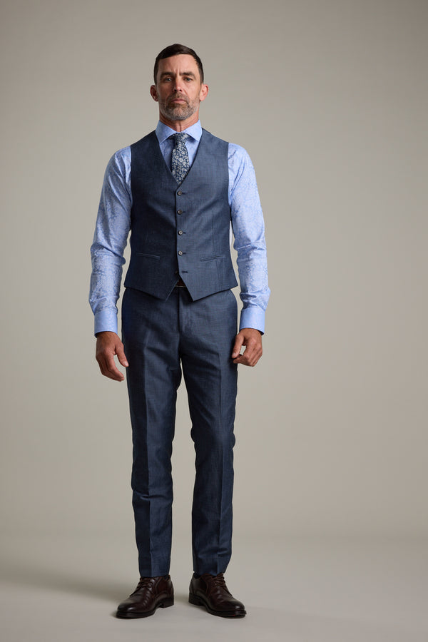 A man with short hair and a beard stands facing forward, wearing the Barkers Charles Texture Waistcoat with an adjustable back buckle, paired with a blue three-piece suit, patterned tie, and brown dress shoes against a plain light gray background.