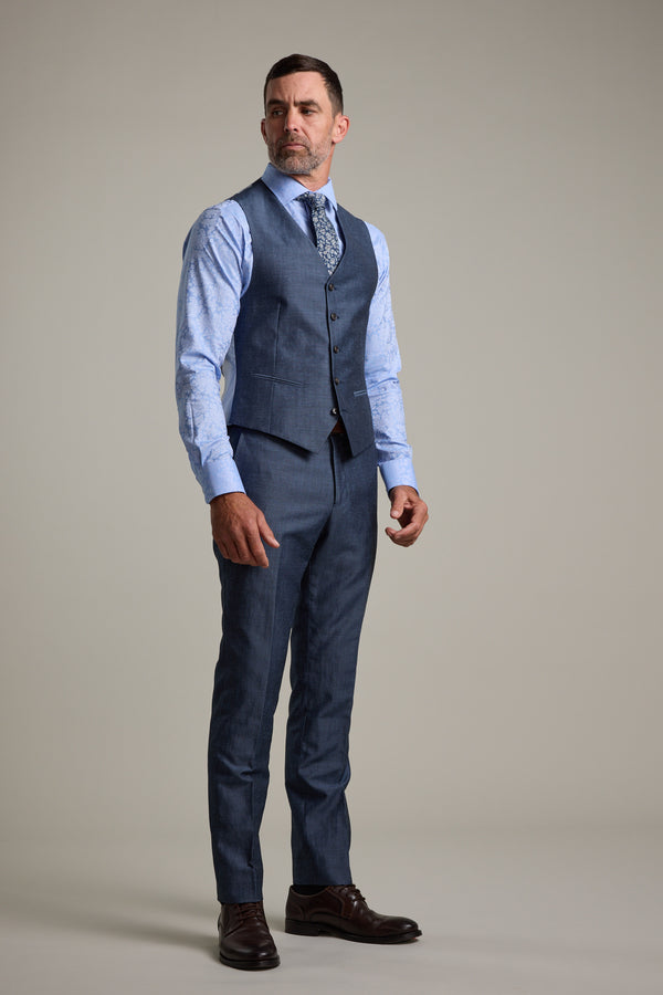 A man stands against a plain background wearing a blue dress shirt, patterned tie, and three-piece suit featuring the Barkers Charles Texture Waistcoat with an adjustable back buckle, finished with dark brown dress shoes. He looks confidently to the side.
