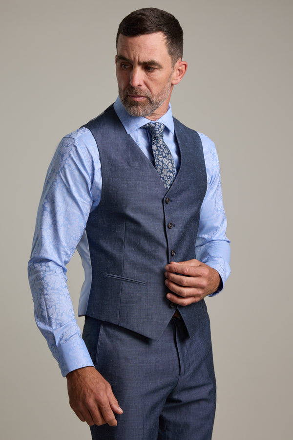 A man with short dark hair and a beard wears the Barkers Charles Texture Waistcoat, part of a blue three-piece suit with a patterned tie, adjusting the vest’s back buckle against a plain background.