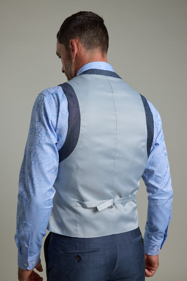 A man is seen from the back wearing the Barkers Charles Texture Waistcoat in light blue linen wool, featuring an adjustable back buckle, layered over a patterned blue shirt and blue trousers against a neutral background.