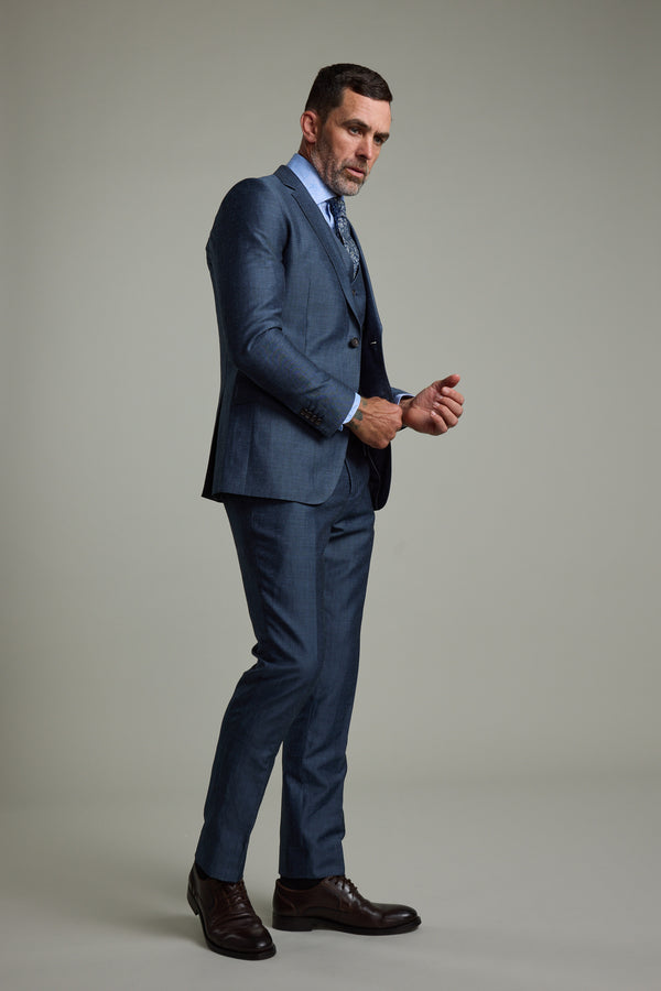 A man poses in a studio wearing the Barkers Charles Texture Suit Jacket with a light blue shirt, patterned tie, and dark brown dress shoes. He looks serious, adjusting his cuff against a plain light gray background.