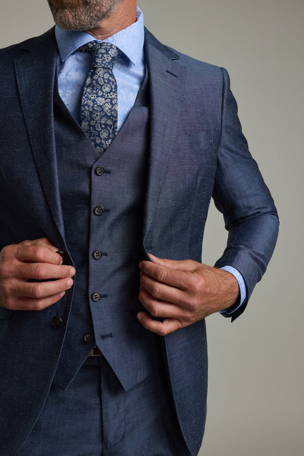 A man in a tailored blue three-piece suit, blue shirt, and patterned tie adjusts his jacket. He wears the Barkers Charles Texture Waistcoat, featuring an adjustable back buckle for a better fit. Cropped from the shoulders down.
