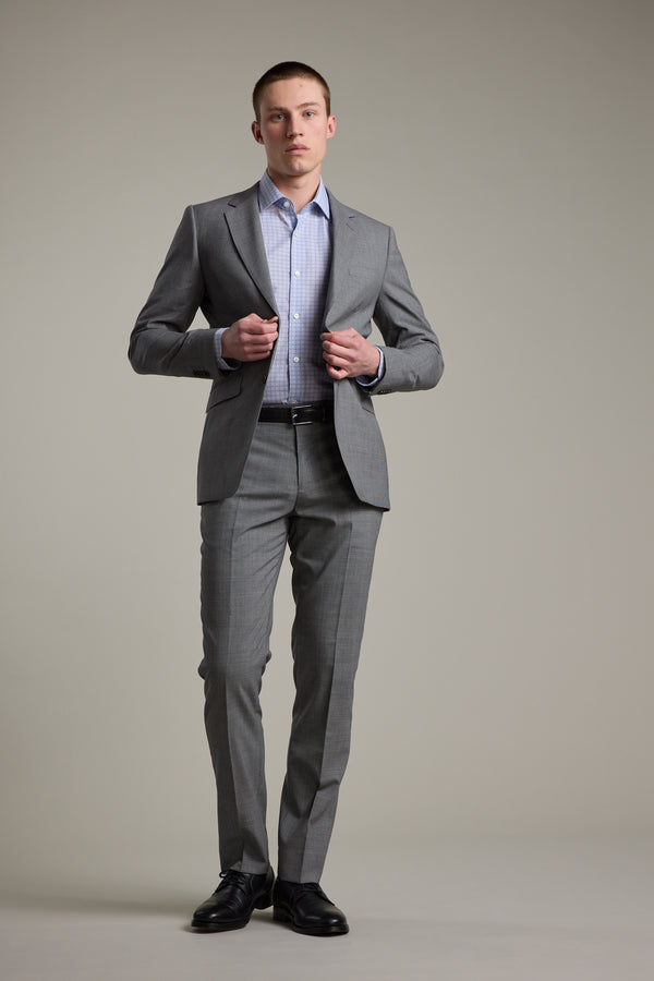 A man stands against a plain background wearing Barkers Henderson Houndstooth Suit Trousers with a light blue dress shirt and black dress shoes as he adjusts his suit jacket.