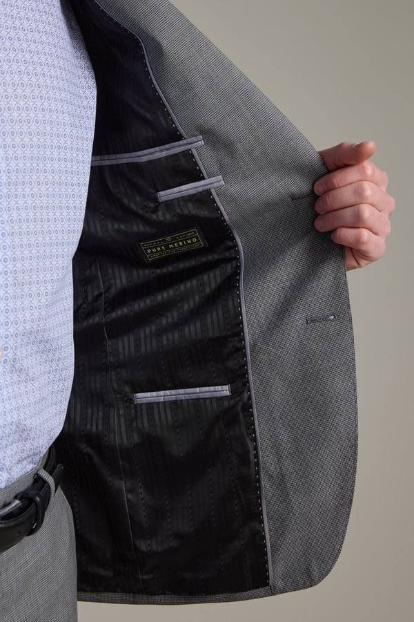 A person holds open a gray Barkers Henderson Houndstooth Suit Jacket, showing the black inner lining and label. They're also wearing gray pants and a light blue patterned shirt; only their lower face and torso are visible.