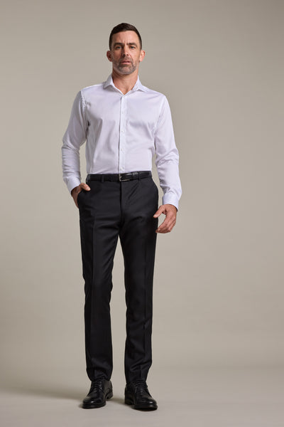 A man stands against a plain background wearing Barkers Mayfair ZQrx Suit Trousers in black, paired with a white button-up shirt, black belt, and black dress shoes. One hand is in his pocket and he has a neutral expression.