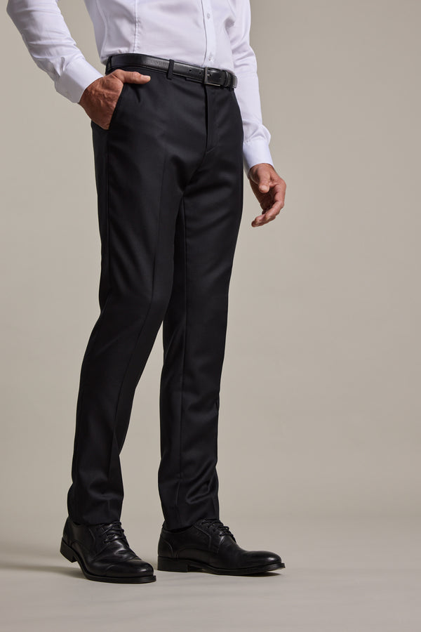 A person wears Barkers Mayfair ZQrx Suit Trouser, a white long-sleeve shirt, black belt, and black dress shoes while standing with one hand in their pocket against a plain background.