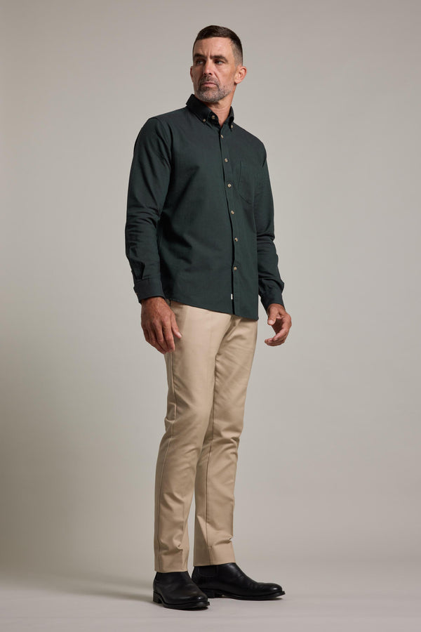 A man with short hair and a trimmed beard stands against a plain background wearing the Lyndon Cotton Pants by Barkers in beige, paired with a dark green button-up shirt and black shoes, looking to the side with a neutral expression.
