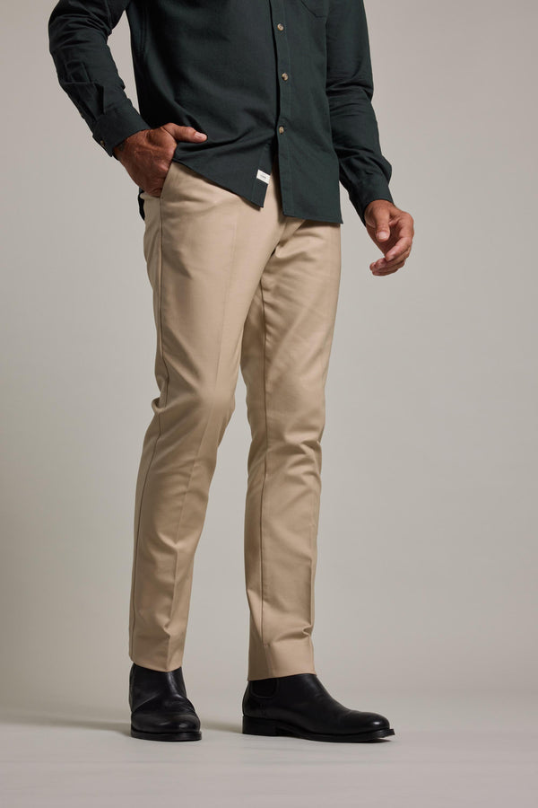 Wearing Barkers Lyndon Cotton Pants in beige, a dark green button-up shirt, and black dress boots, a person stands with one hand in their pocket against a plain background, exemplifying a smart casual style.