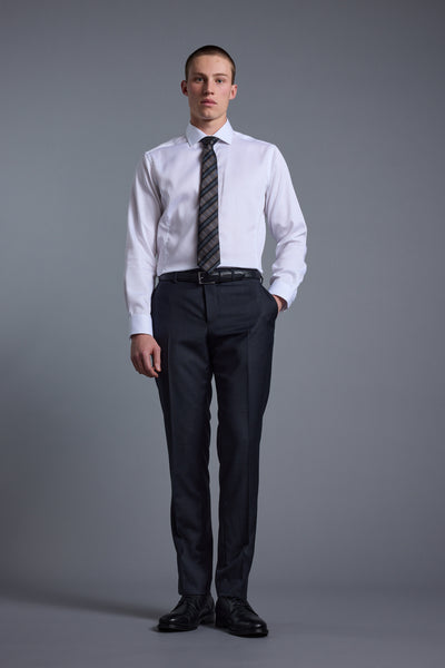 A man stands against a plain gray background, wearing a white dress shirt, patterned tie, black shoes, and Barkers Mayfair ZQ Suit Trouser. One hand is in his pocket and he has a neutral expression.