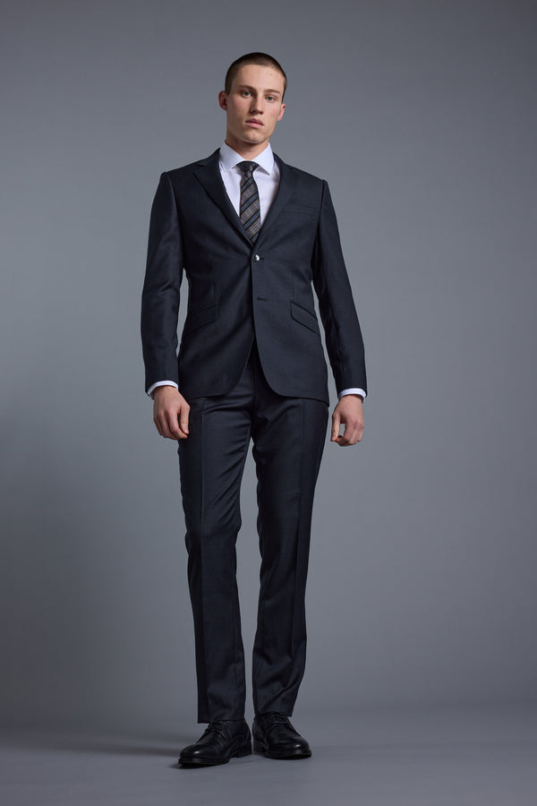 A man with short hair and a neutral expression stands against a gray background wearing the Barkers Mayfair ZQ Suit Jacket, crafted from breathable ZQrx Merino Wool. His arms are relaxed at his sides.