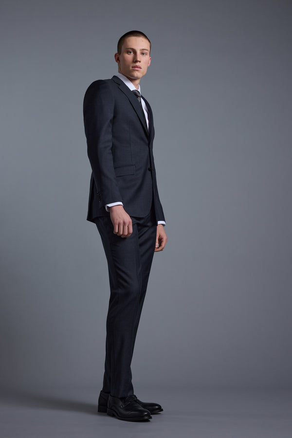 A young man in Barkers' Mayfair ZQ Suit Trouser, paired with a dark three-piece suit, white dress shirt, and tie, stands against a plain gray background, gazing slightly left with a serious expression.