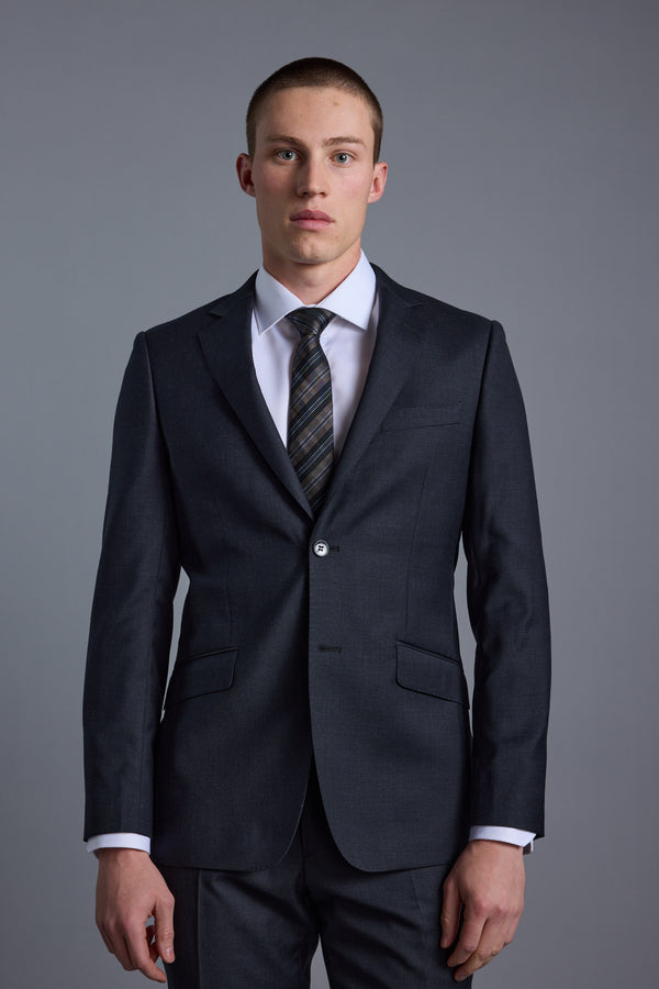 A young man with short hair and a neutral expression stands facing forward against a plain gray background, wearing the Barkers Mayfair ZQ Suit Jacket made from ZQ Merino Wool.