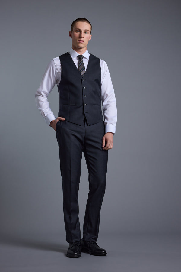 A man stands against a gray background, wearing a dark three-piece suit featuring the Barkers Mayfair ZQ Waistcoat made from ZQ-certified New Zealand merino wool. He has one hand in his pocket and a neutral expression.