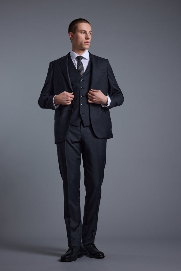 A man in a dark New Zealand merino wool three-piece suit, white shirt, and Barkers Mayfair ZQ Waistcoat stands against a plain gray background, looking to his left while adjusting his jacket.