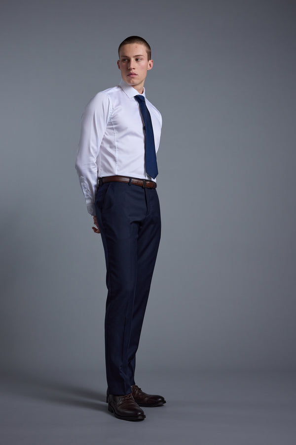 A young man stands against a gray background, wearing Barkers’ Mayfair ZQ Suit Trouser, a white dress shirt, navy blue tie, brown belt, and brown shoes. He looks to the side with his hands behind his back.