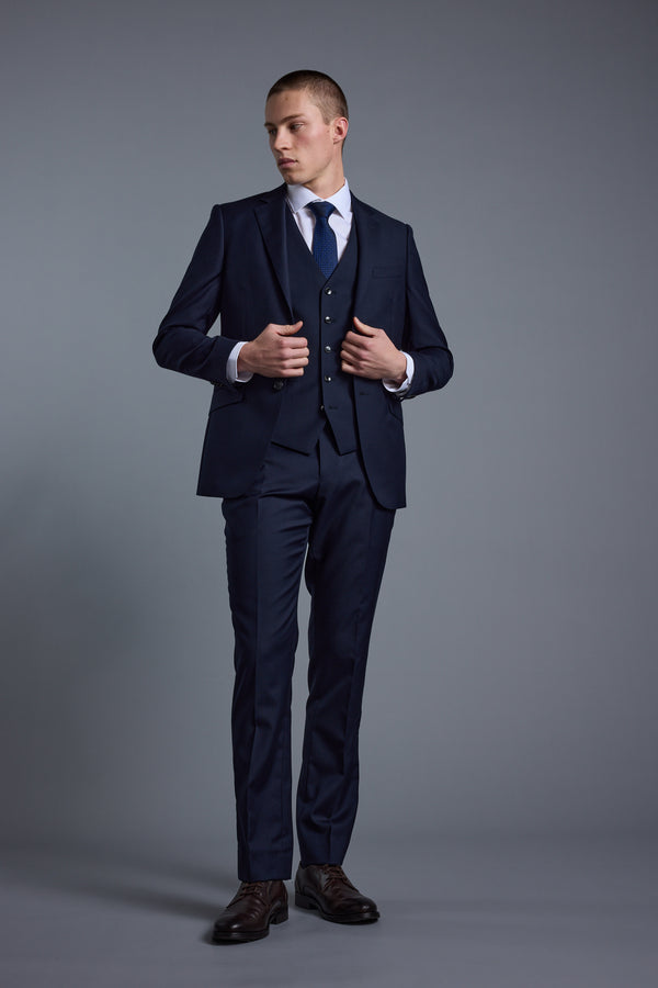 A man in a dark blue Barkers Mayfair ZQ Suit Jacket, light blue shirt, and tie stands against a plain gray background, looking to his left while adjusting his vest.