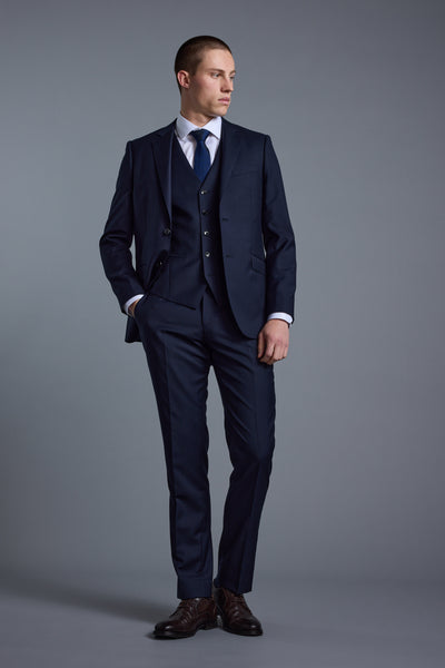 A man wearing a Barkers Mayfair ZQ Waistcoat in navy blue New Zealand merino wool, paired with a matching suit, white shirt, and blue tie stands with one hand in his pocket, looking to the side against a plain gray background.