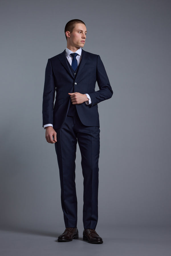 A man stands against a plain gray background wearing Barkers' Mayfair ZQ Suit Trouser in dark blue New Zealand merino wool, paired with a matching jacket, white shirt, and navy tie. He adjusts his jacket while looking slightly to the side.